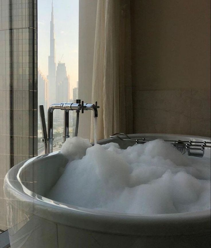 a bathtub filled with foam and water next to a large window overlooking a city