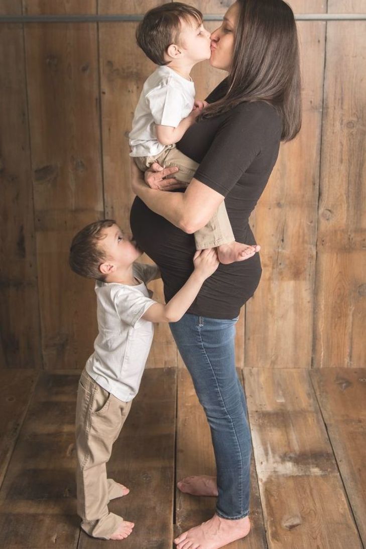 a woman holding a baby and kissing it's face while standing in front of a wooden wall