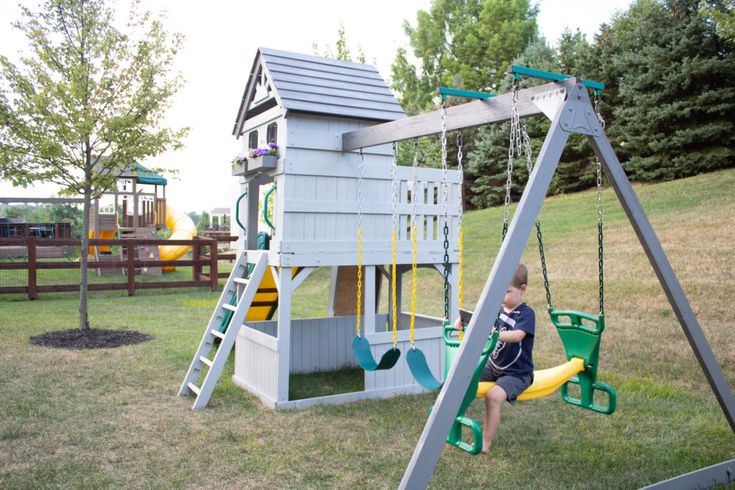 a little boy sitting on a swing set in the grass next to a play house