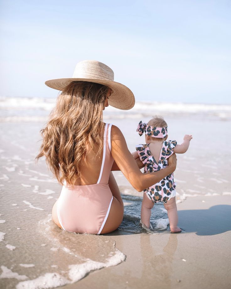 a woman in a bathing suit and hat sitting on the beach next to a baby