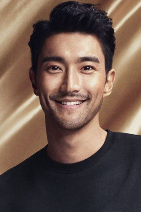 a smiling man in a black t - shirt with a gold satin background behind him