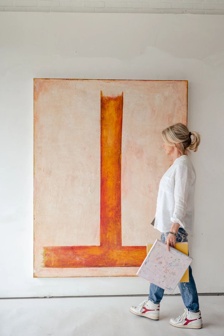 a woman in white shirt and jeans walking by large painting with orange line on it