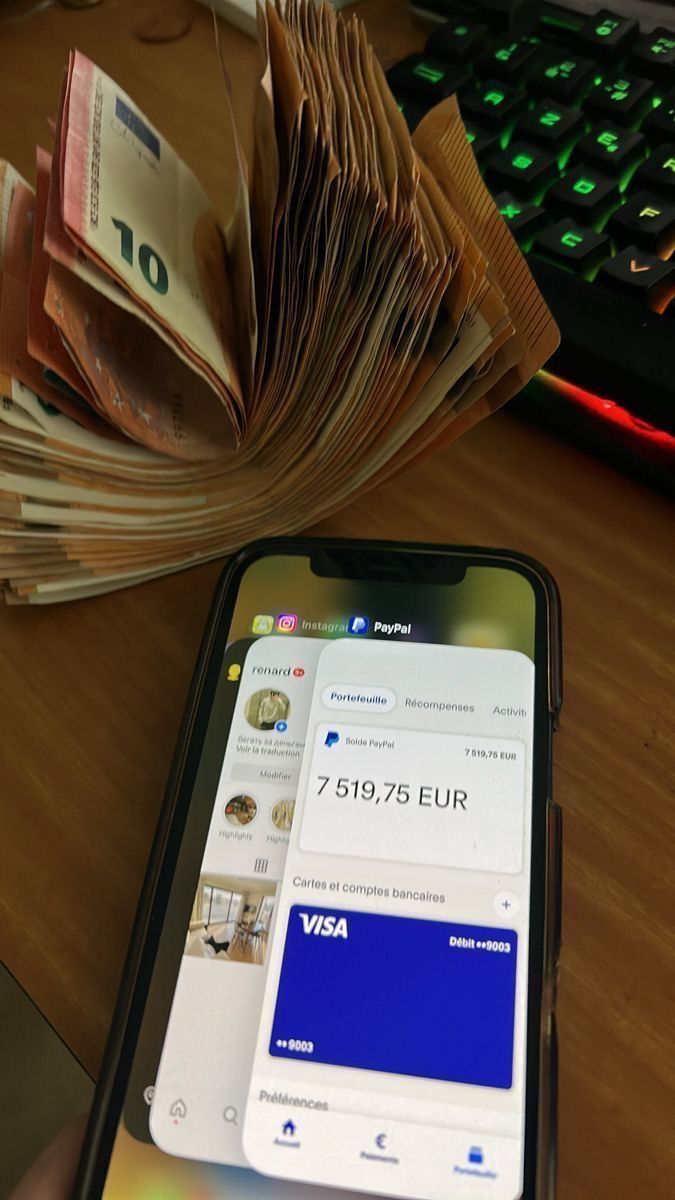 a person holding a cell phone next to a stack of money on a desk with a keyboard