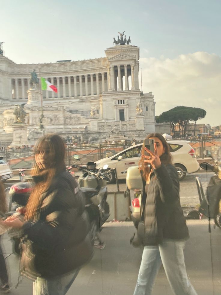 two women are walking down the street with their cell phones up to their ears and one is taking a photo