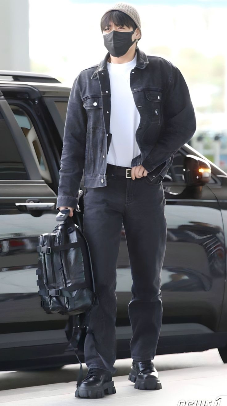 a man wearing a face mask and carrying a suitcase in front of a car at an airport