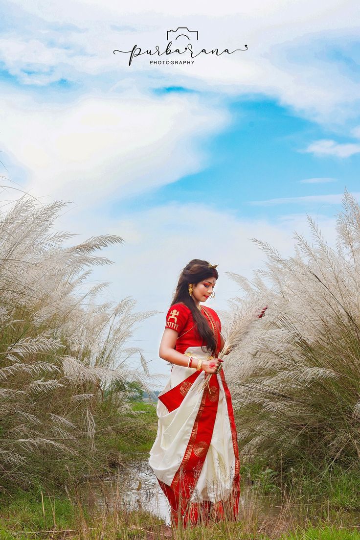 a woman in a red and white sari is walking through tall grass with her hand on her hip