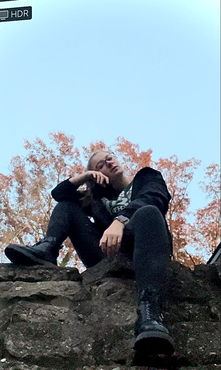 a man sitting on top of a large rock next to a leaf filled tree and talking on a cell phone