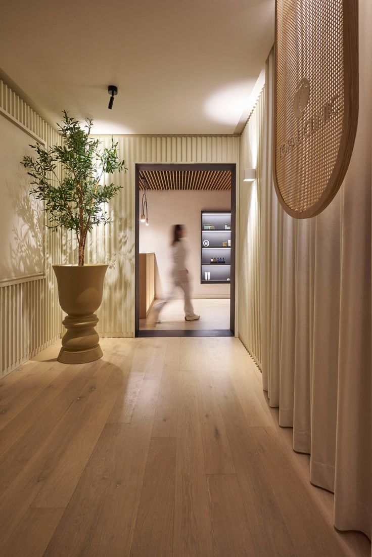 a person walking down a hallway with a potted plant in the foreground and an open door leading to another room