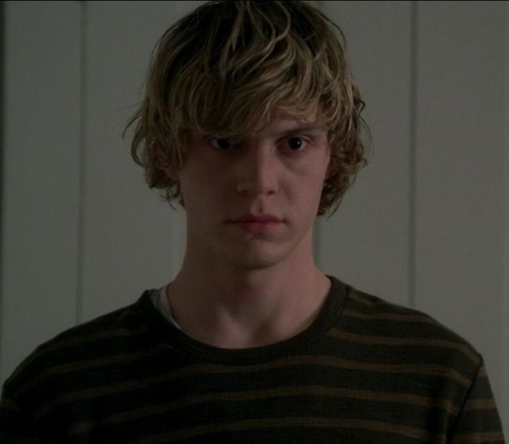 a young man standing in front of a white wall wearing a black and brown striped shirt