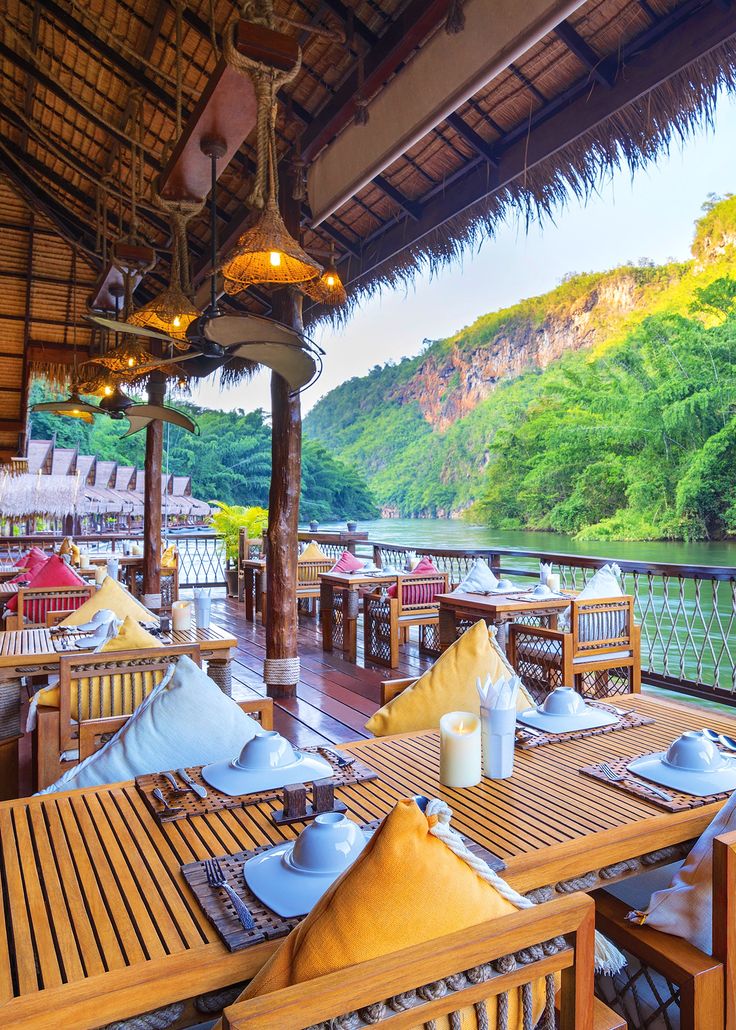 an outdoor dining area with wooden tables and chairs overlooking the river in front of mountains