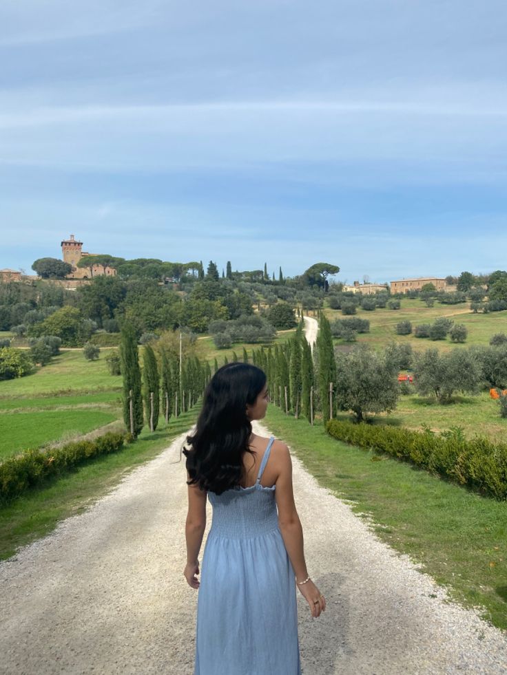 a woman in a blue dress walking down a dirt road with trees on either side