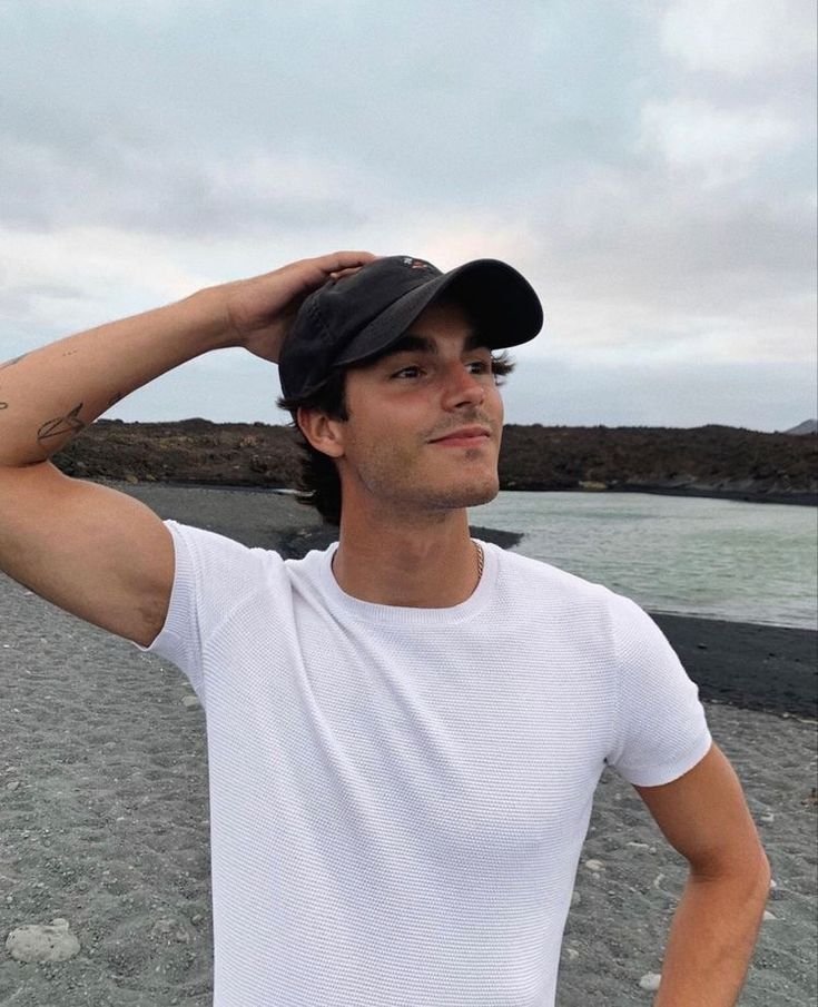 a man in white shirt and black hat standing on beach next to body of water