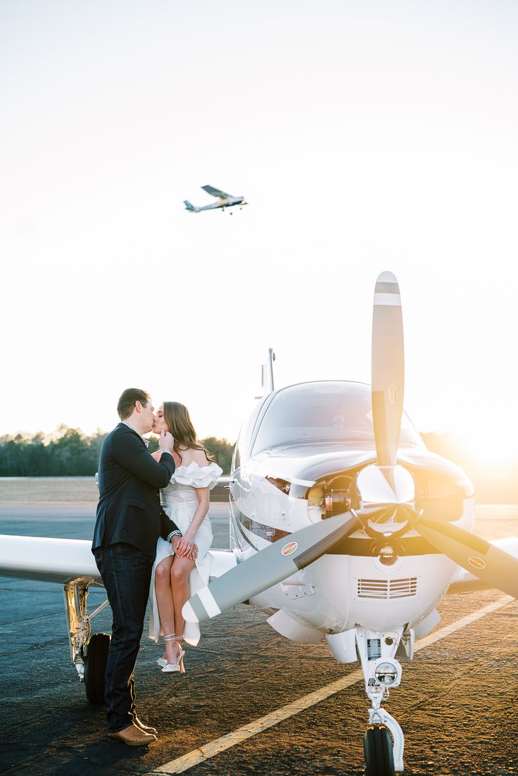 a bride and groom kissing in front of an airplane on the tarmac at sunset