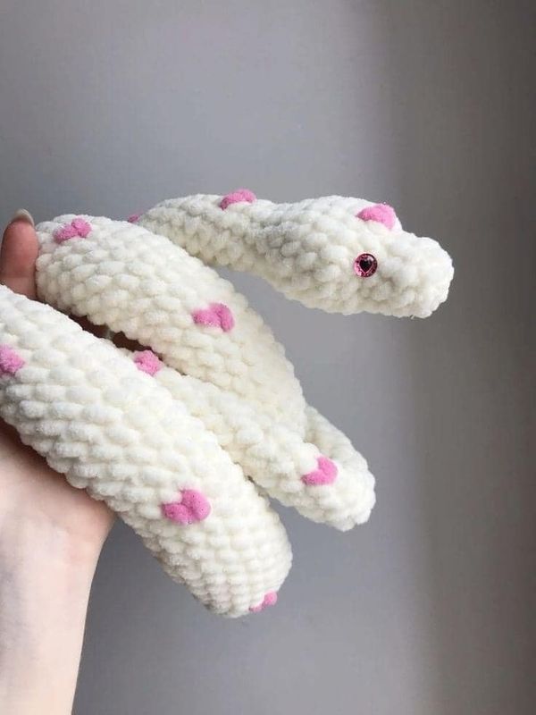 a hand holding two white knitted snakes with pink hearts