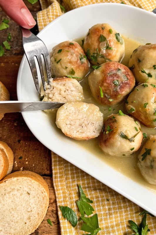 a white bowl filled with meatballs and gravy next to bread slices on a wooden table