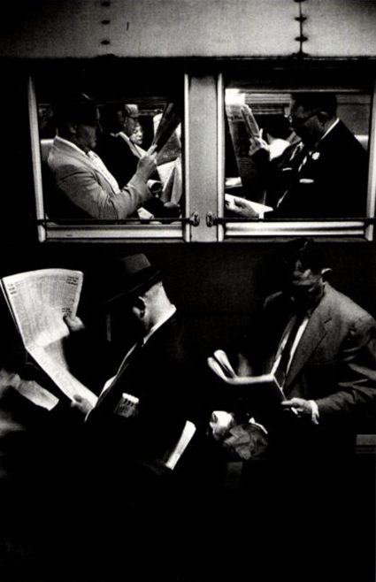 black and white photograph of two men sitting on a subway car reading newspapers while another man looks out the window