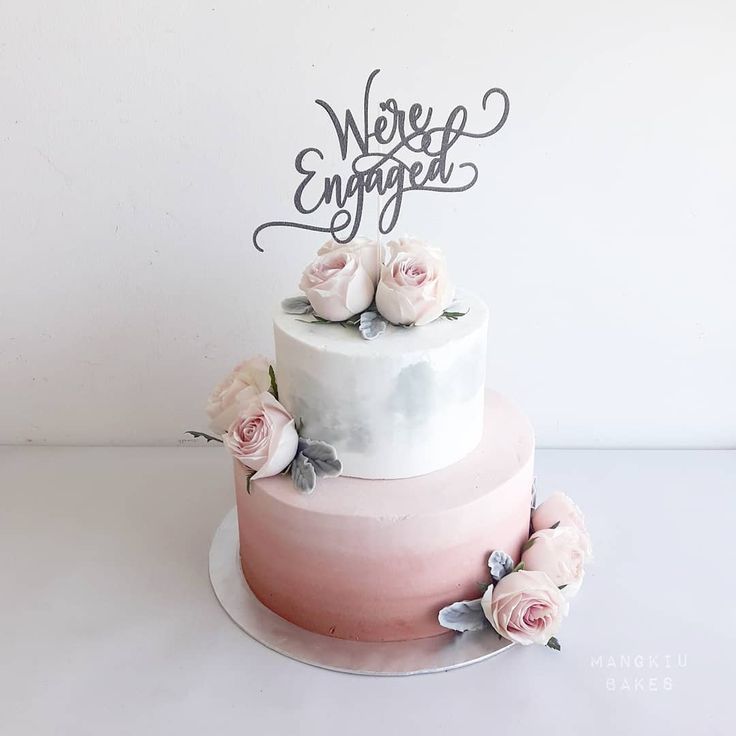 a three tiered cake with flowers on top that says we're engaged written in cursive letters