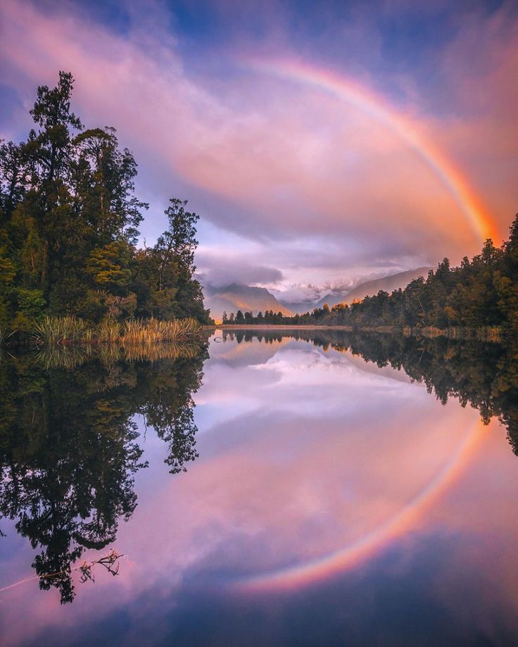 a rainbow in the sky over a lake with trees and mountains reflected in the water