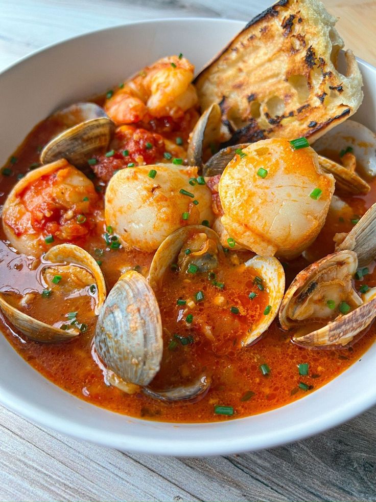 a white bowl filled with seafood and clams on top of a wooden table next to bread