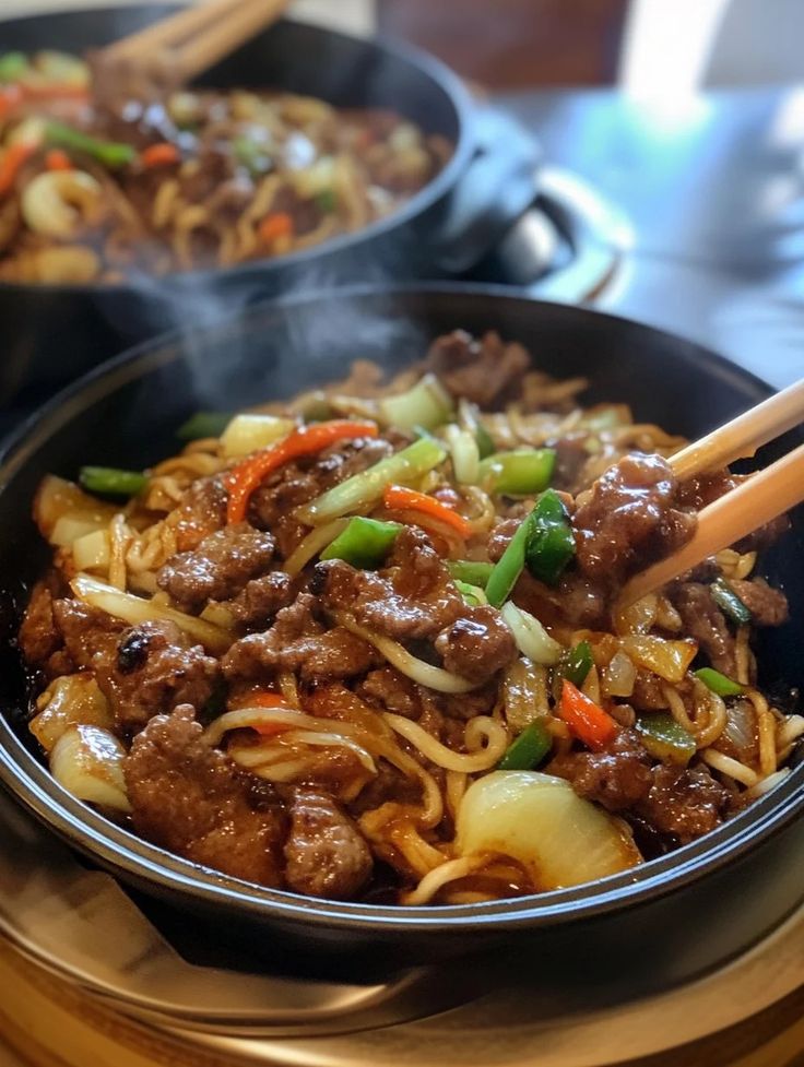 two pans filled with beef and noodles on top of a wooden table next to chopsticks