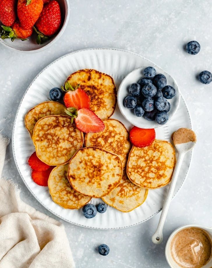 pancakes with strawberries and blueberries are on a plate next to a bowl of fruit