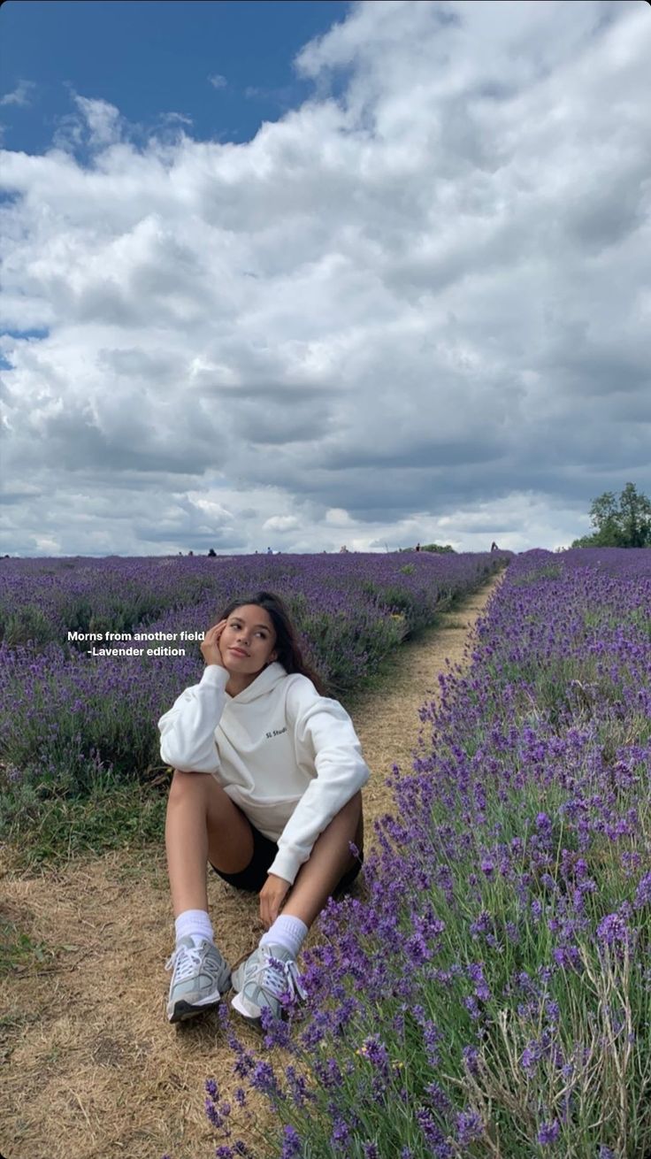 a woman sitting on the side of a road next to a field of purple flowers