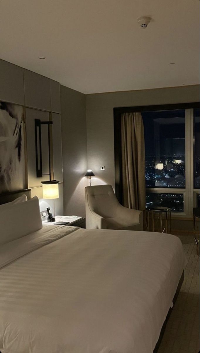 a hotel room with a bed, chair and large window overlooking the city at night