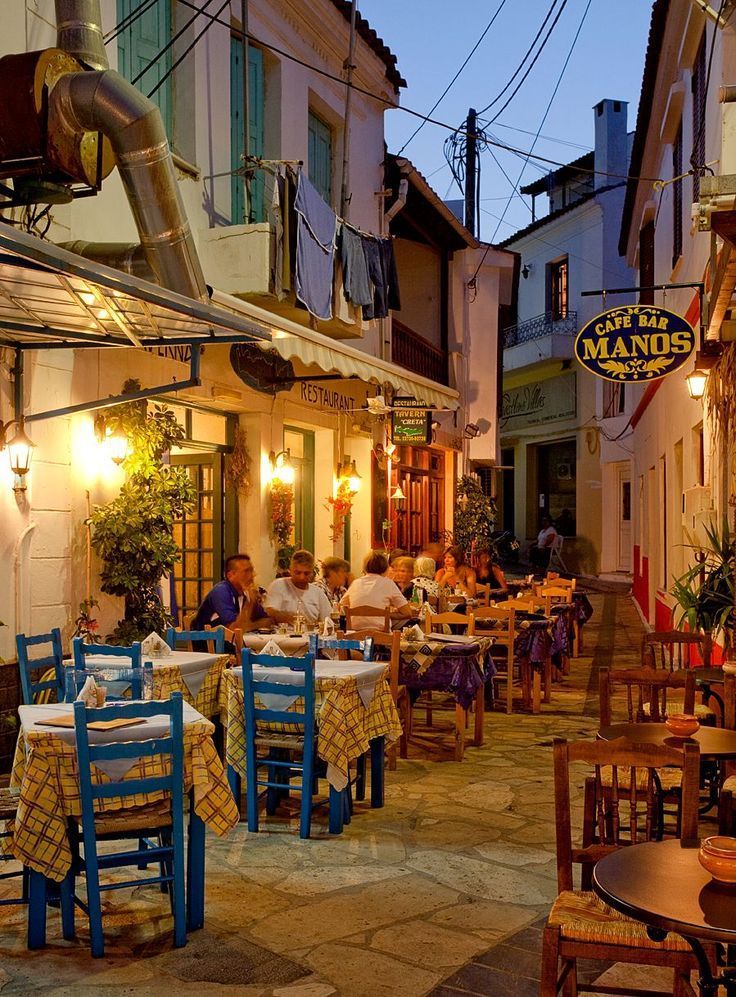 people are sitting at tables in an alleyway with many restaurants and bars on either side