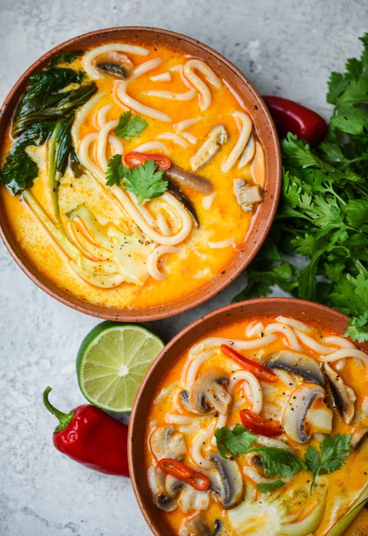 two bowls filled with noodles and vegetables on top of a table next to lime wedges