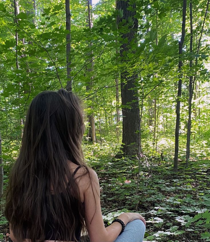 a woman sitting in the woods with her back to the camera and looking at trees