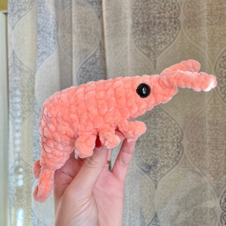 a pink stuffed animal that is in someone's hand with a curtain behind it