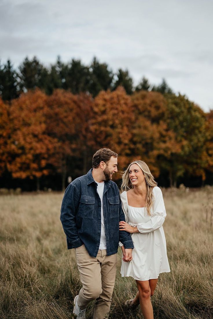 a man and woman walking through tall grass in an open field with trees behind them