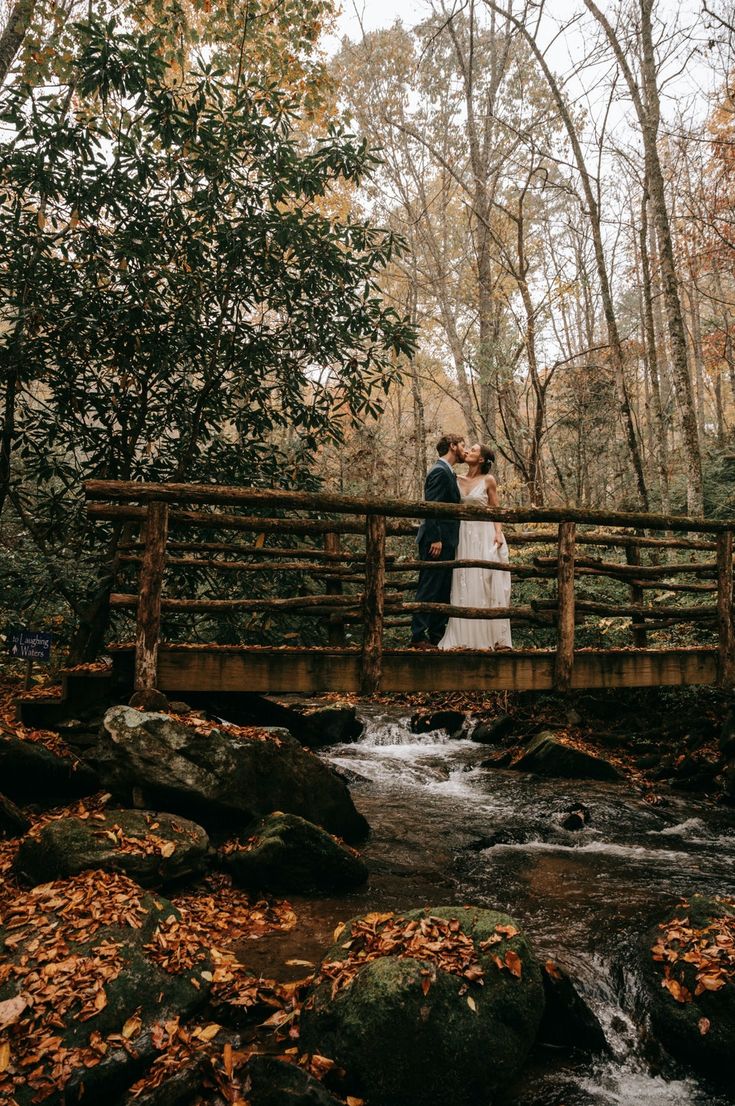 a bride and groom are standing on a bridge over a stream in the woods with fall foliage