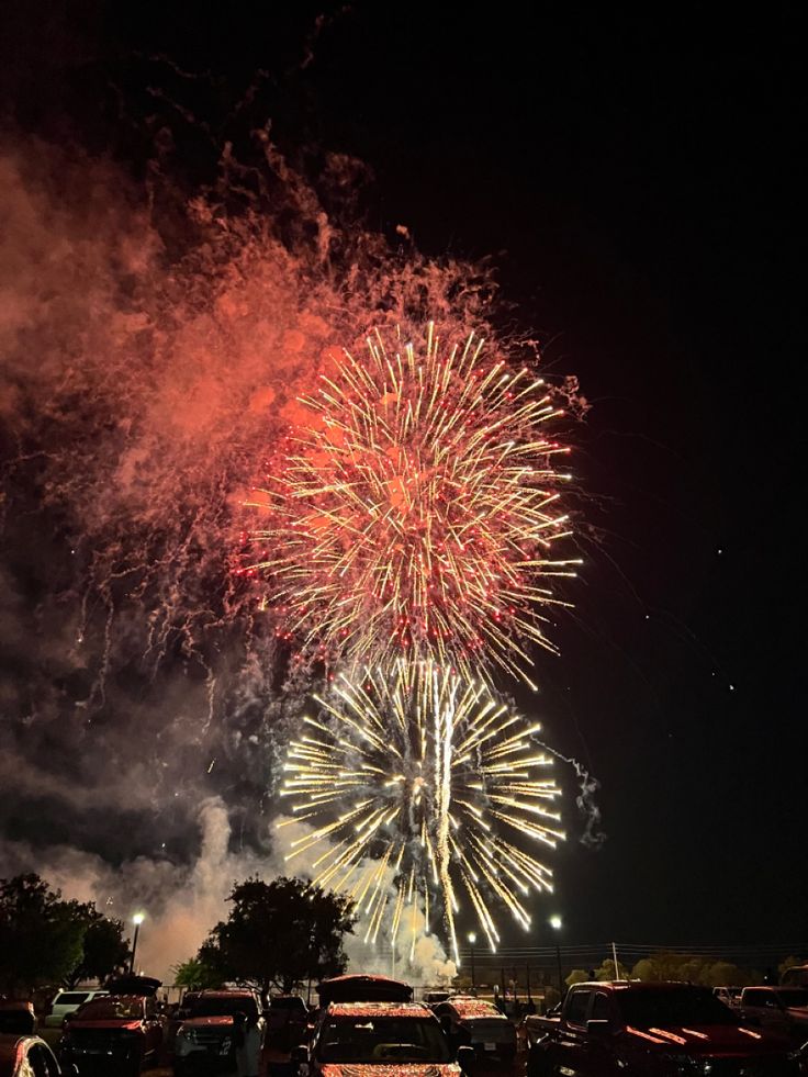 fireworks are lit up in the night sky above parked cars and people standing around them