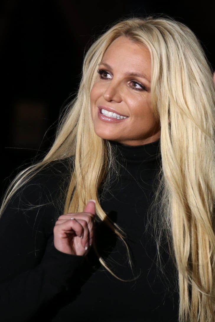 a woman with long blonde hair and black top smiling at the camera while standing in front of a dark background