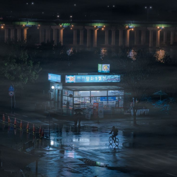 a person riding a bike in the rain at night near a building with lights on