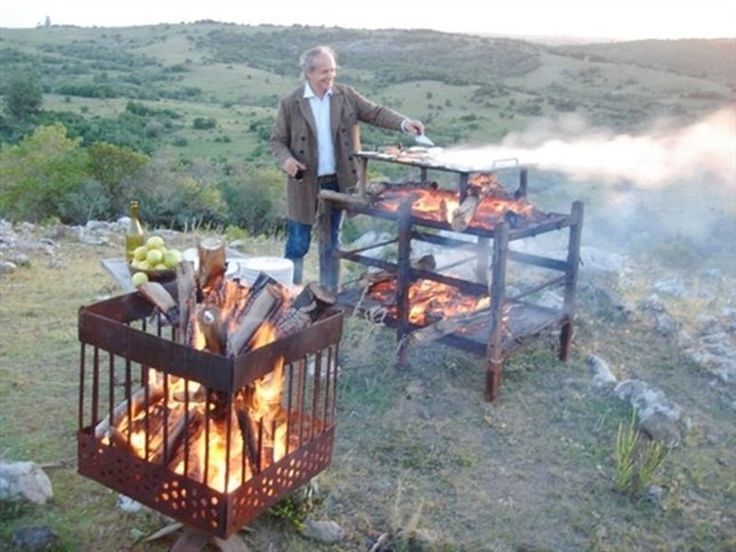 a man standing next to a grill on top of a grass covered field with flames