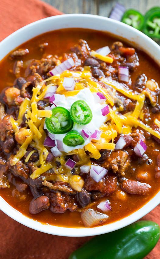 a white bowl filled with chili and cheese on top of a wooden table next to green peppers