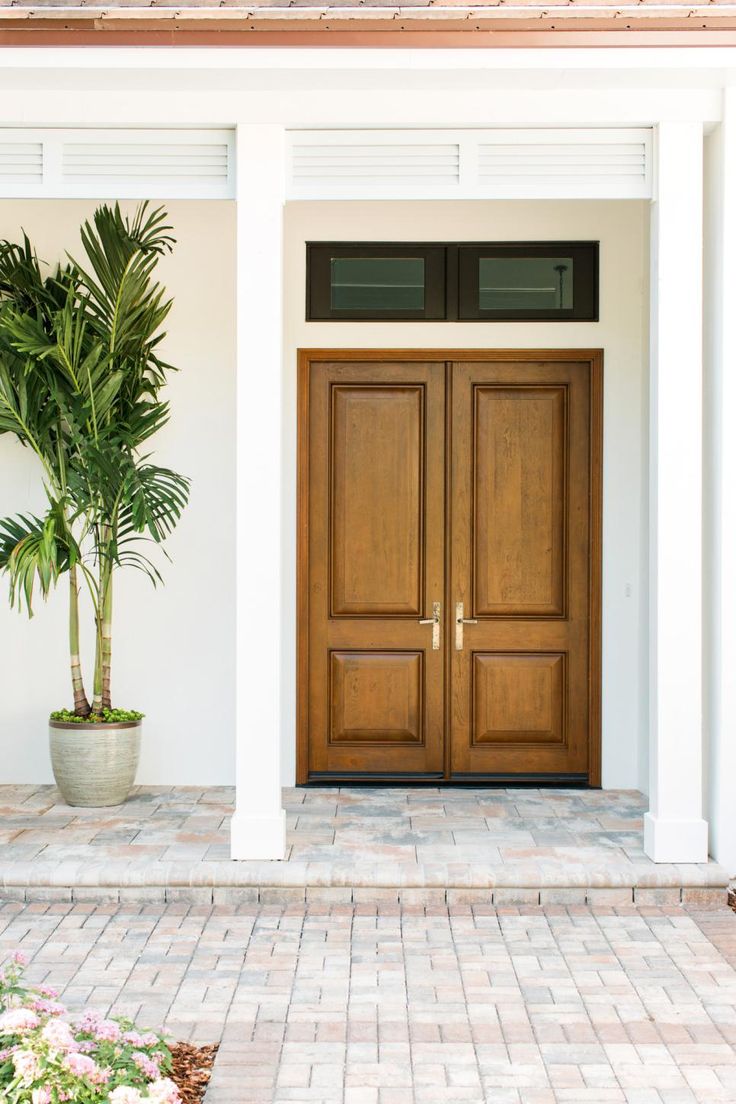 two wooden doors on the front of a white house with brick walkway and potted palm tree