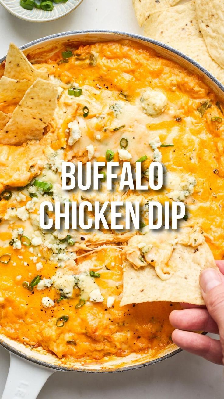 a hand holding a tortilla chip over a skillet filled with buffalo chicken dip