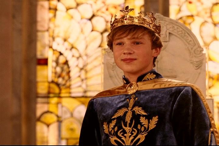 a young man dressed in medieval clothing and wearing a tiara, standing next to a stained glass window