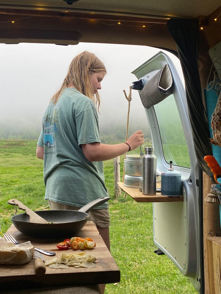 a woman cooking food out of an open camper door on a foggy day