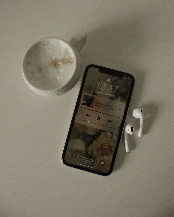 an iphone with ear buds next to it and a cup on the table in front of it