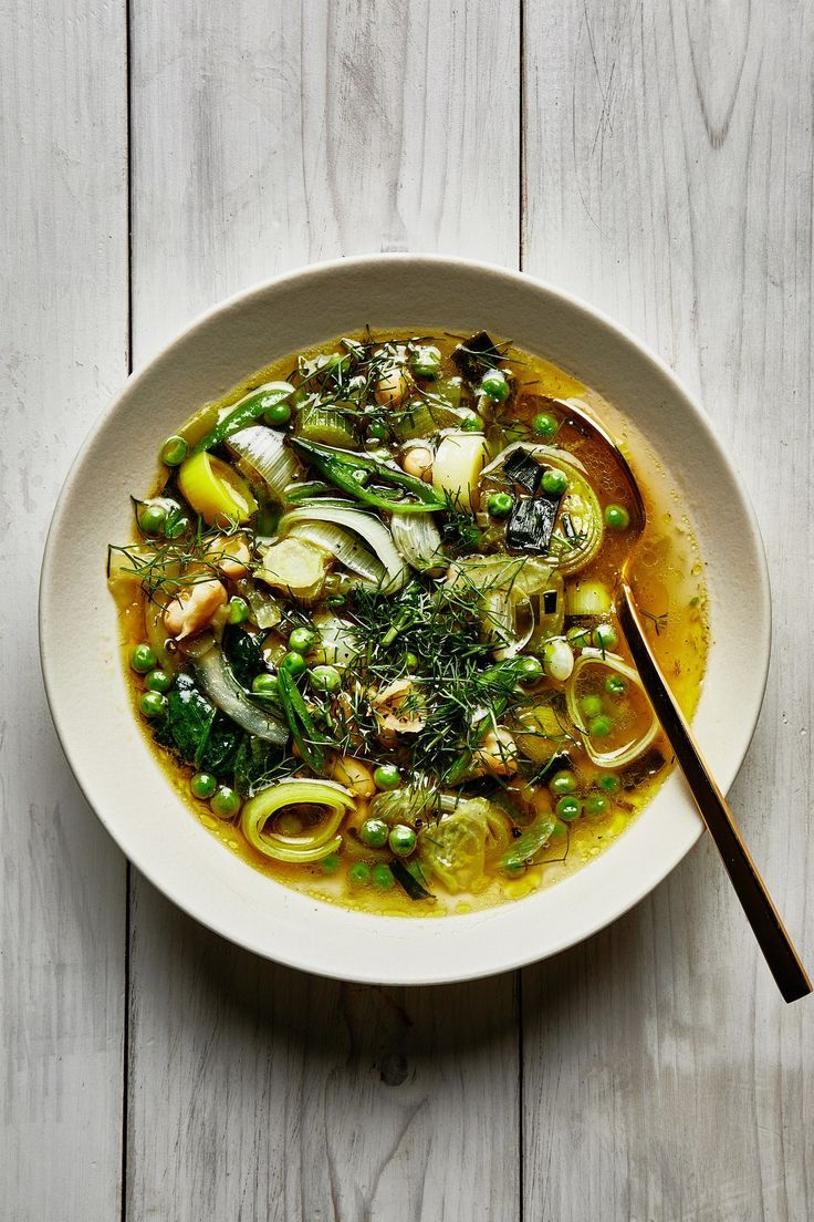 a white bowl filled with soup and vegetables on top of a wooden table next to a spoon