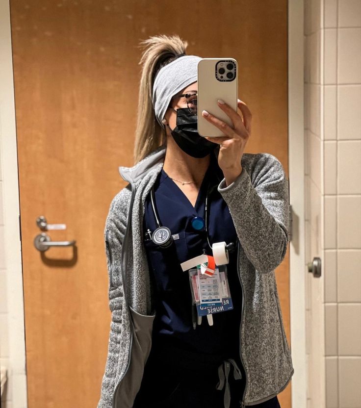 a woman in scrubs takes a selfie with her phone while wearing a mask