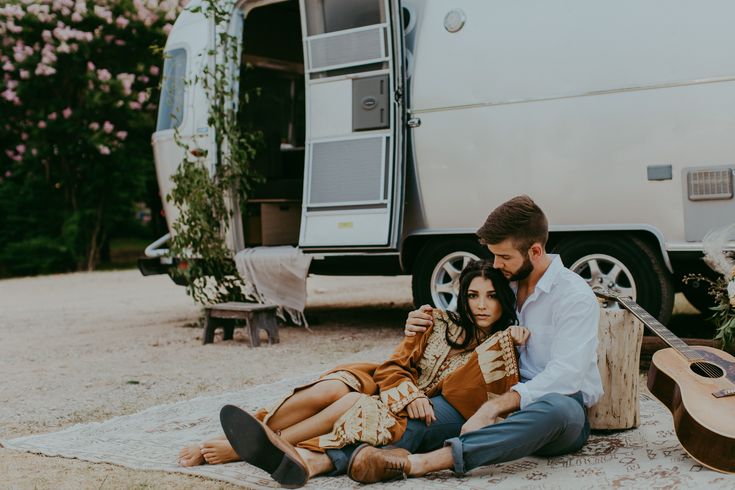 a man and woman sitting on the ground next to a camper trailer with their guitar