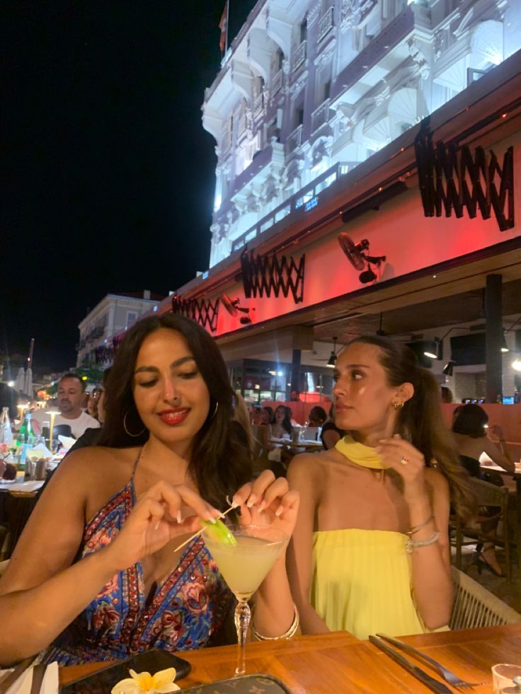 two women sitting at a table with food and drinks in front of them on a city street