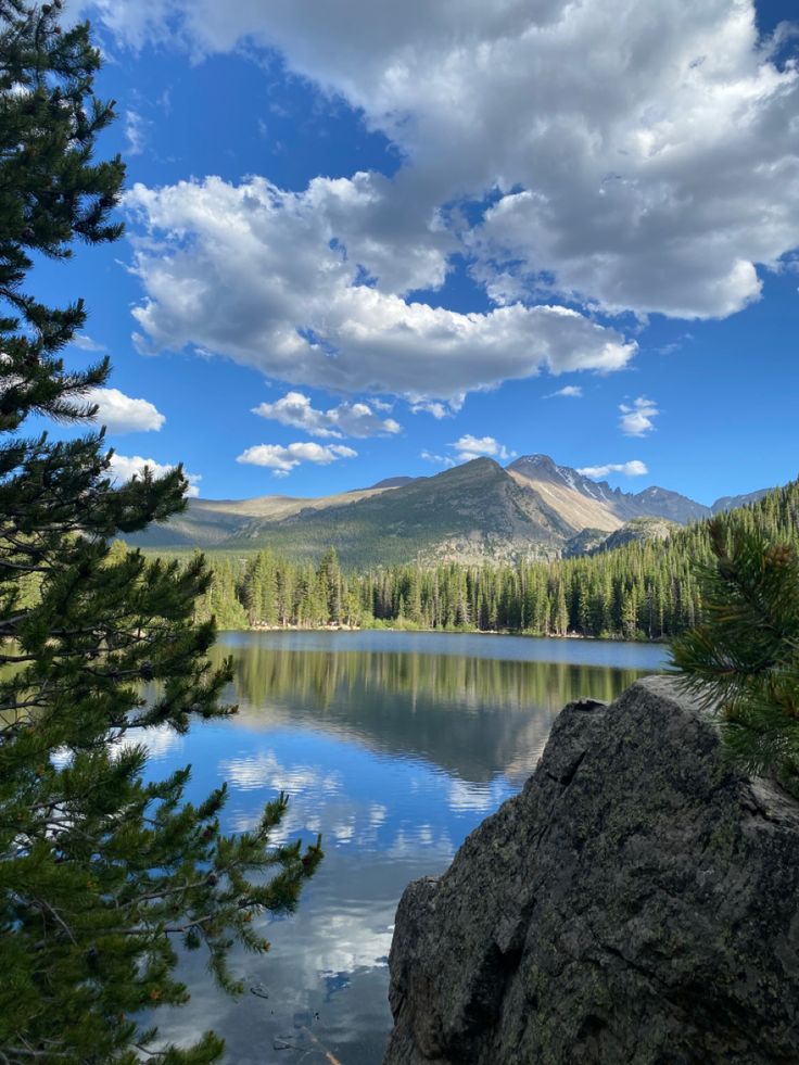 a lake surrounded by trees and mountains under a cloudy blue sky with white fluffy clouds