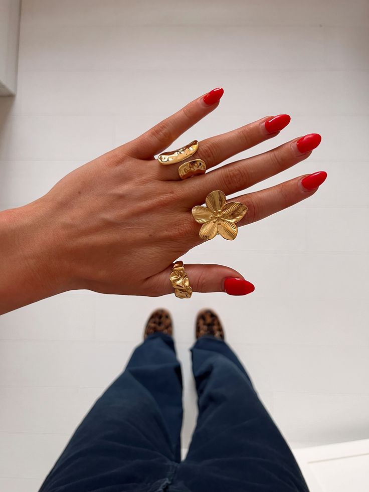 A close-up of a hand with red nail polish, wearing multiple gold rings including a flower ring, against a background of pants and leopard print shoes Jewelry Stacks Gold, Summer Jewelry Stack, Chunky Ring Stack, Stacked Rings Aesthetic, Pretty Stacks, Maximalist Jewelry, Chunky Gold Jewelry, Jewelry Stack, Photography Styling