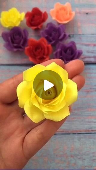 a hand holding a yellow flower in front of colorful paper flowers on a wooden table